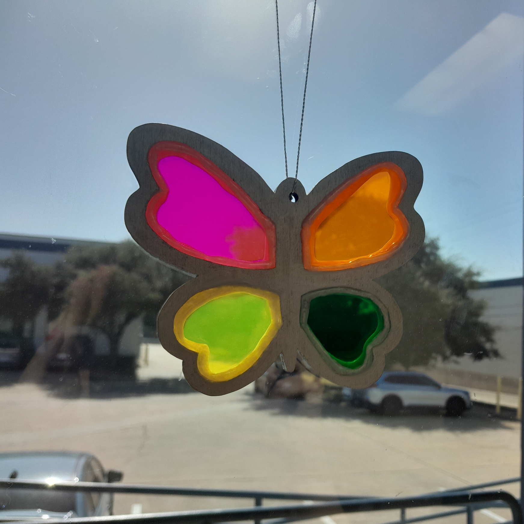 Butterfly-shaped wooden suncatcher with pink, orange, yellow, and green acrylic wings hanging in a window.